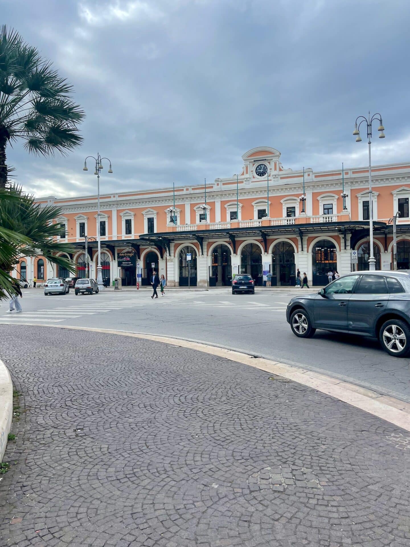 A view of the Bari train station, featuring a large, peach-colored building with white trim and a clock on the top. The station has a grand, historic look with arched entrances and street lamps in front. Cars and pedestrians are visible in the foreground.