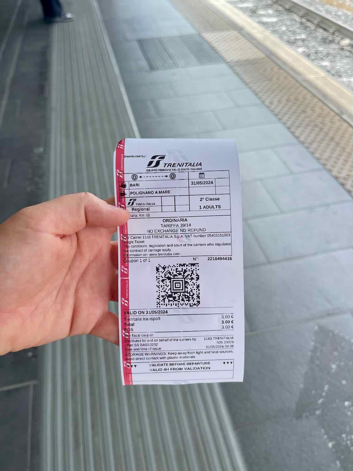 A close-up photo of a Trenitalia train ticket held by a person at a train station in Bari, Italy. The ticket indicates a journey from Bari to Polignano a Mare on 31/05/2024 in 2nd class. It includes a QR code for validation and a fare of 3.00 Euros.