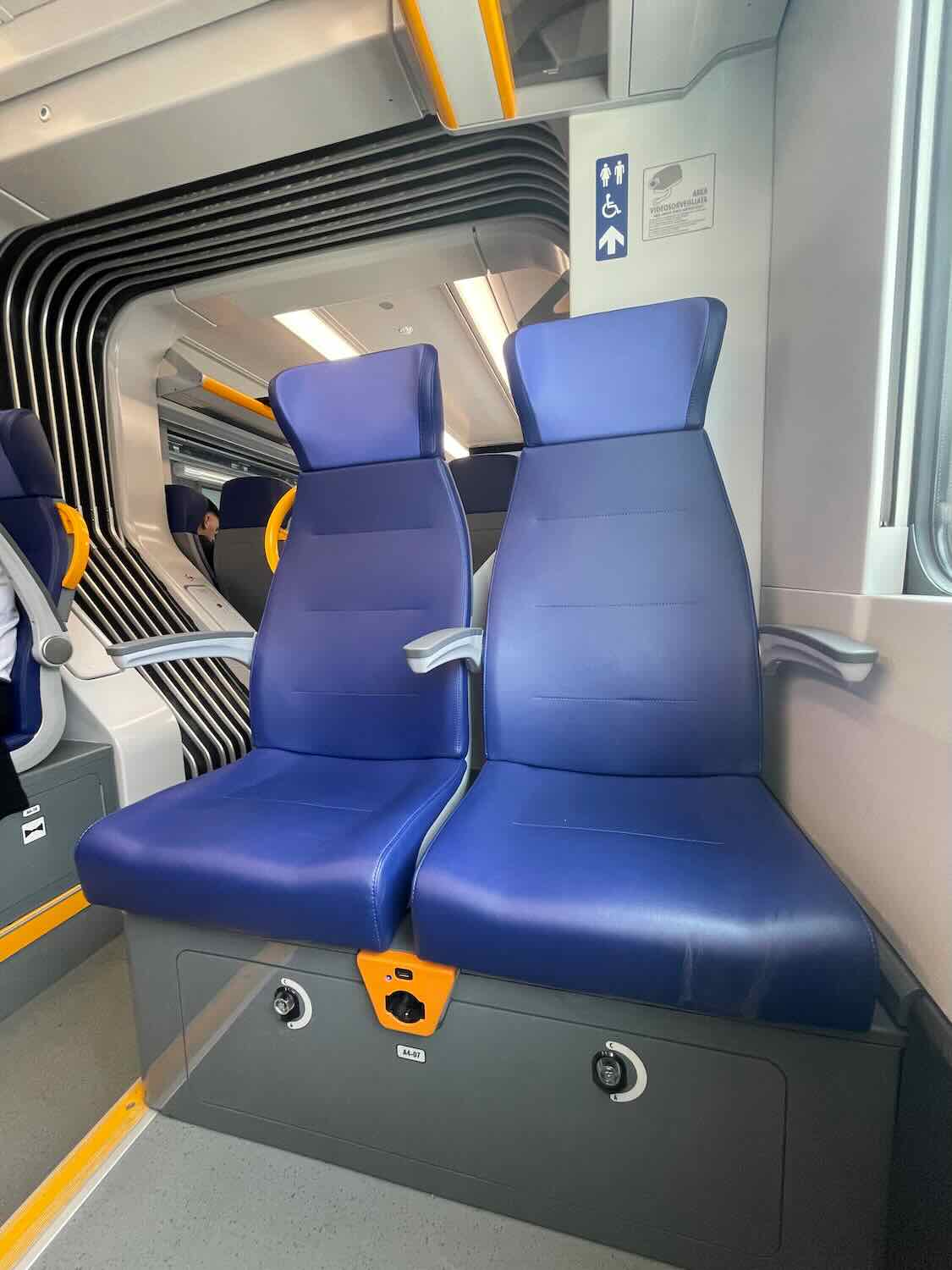 A photo of two blue train seats inside a Trenitalia train in Bari, Italy. The seats are positioned side by side with a modern design, featuring armrests and a small orange panel with a power outlet at the base. The interior of the train is clean and well-lit, with signage indicating the location of accessible facilities.