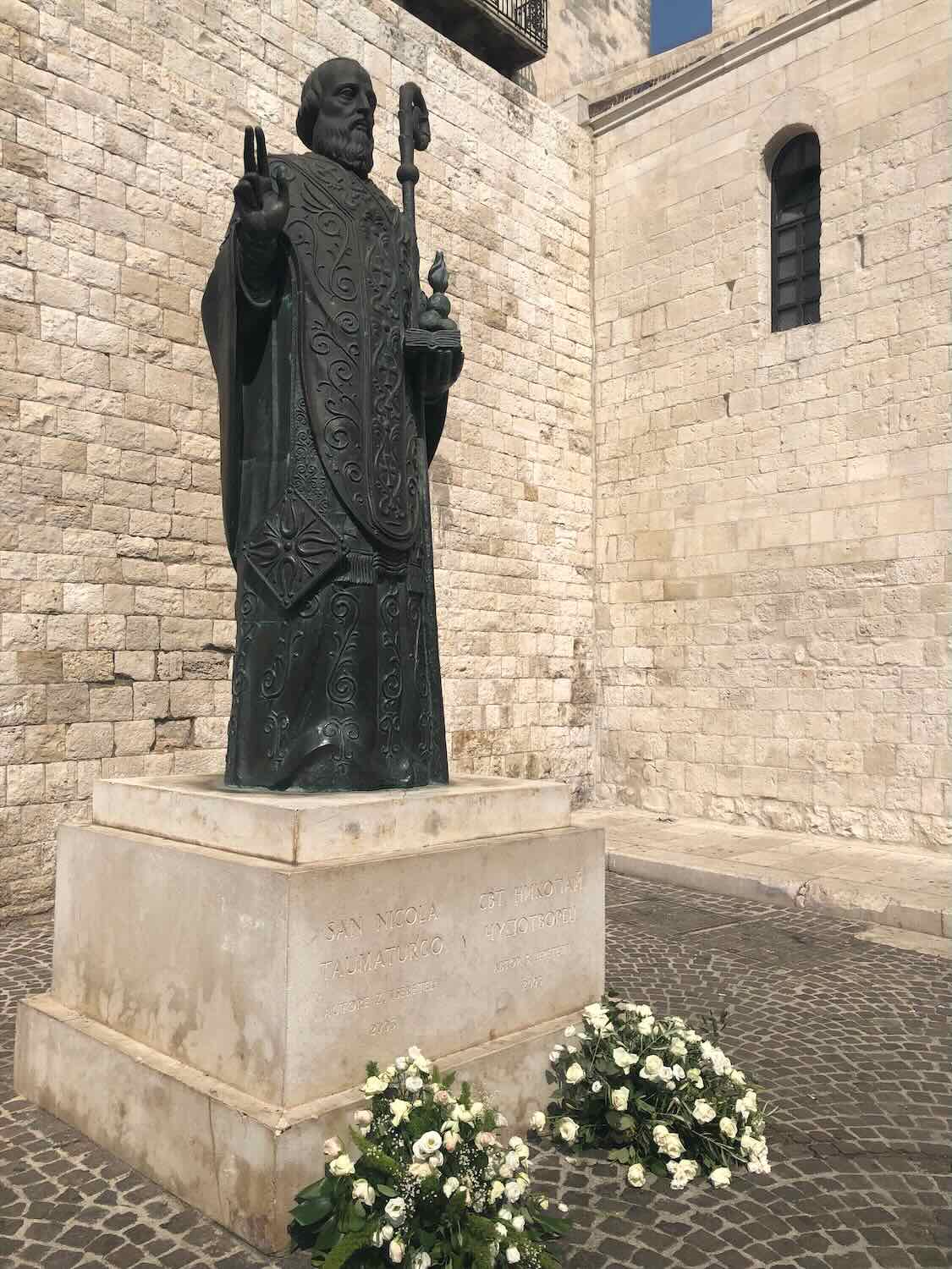 A bronze statue of Saint Nicholas holding a staff and giving a blessing gesture, set on a stone pedestal with inscriptions. The statue is surrounded by white flowers and is positioned against a backdrop of old stone walls.