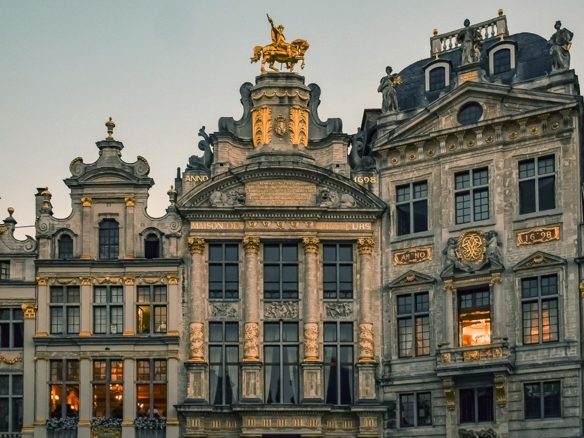 A detailed view of the ornate facades of historical buildings in Brussels' Grand Place. The central building features a golden statue on top, elaborate carvings, and multiple windows with golden accents.