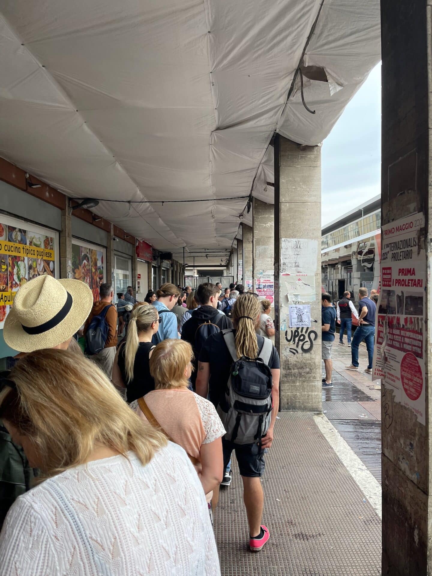 A long queue of people waiting under a covered walkway in Bari, Italy. The crowd consists of tourists and locals, some with backpacks, waiting patiently. The surrounding area has posters and advertisements on the walls.