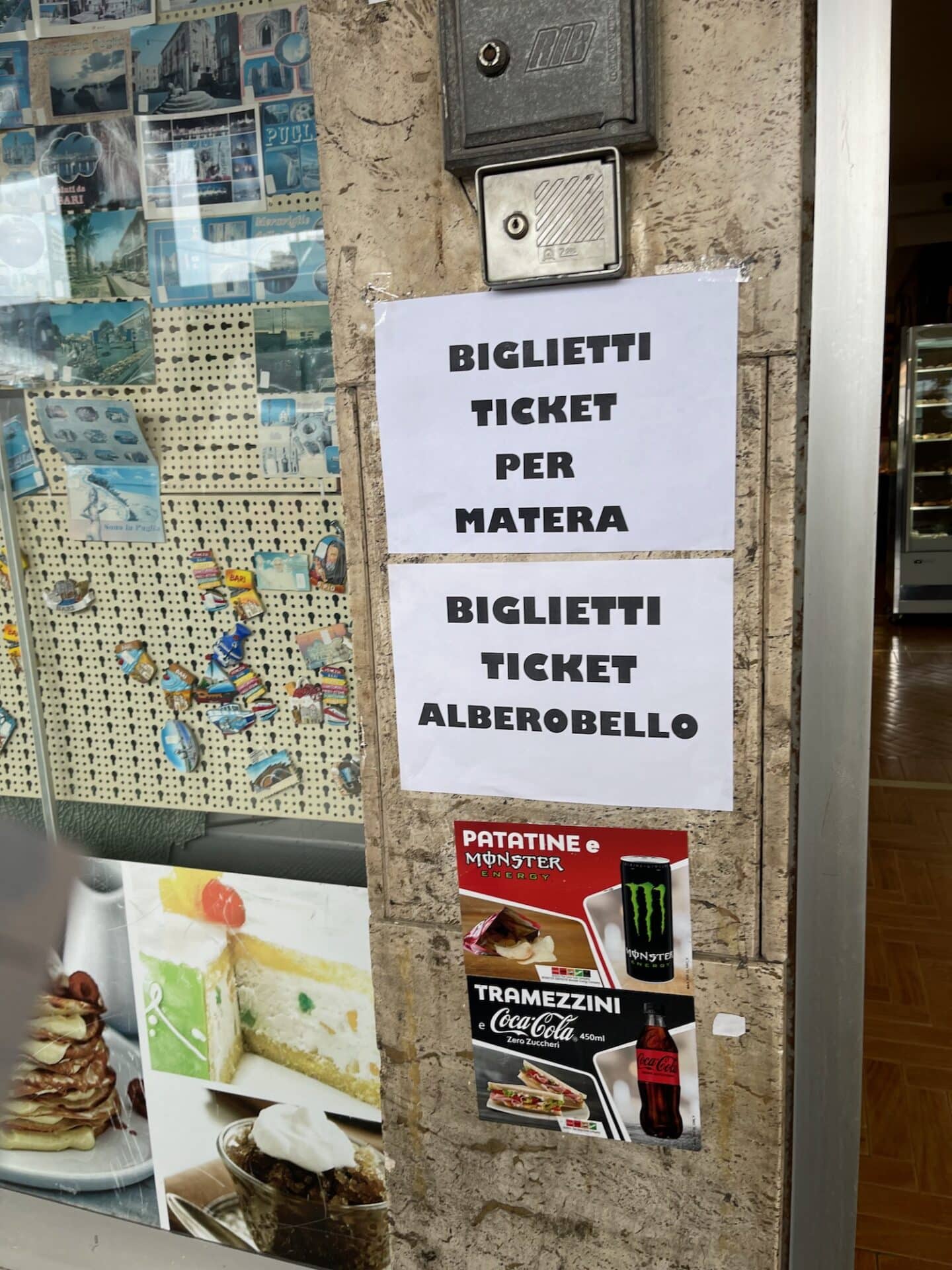 A sign on a wall in Bari, Italy, advertising tickets for Matera and Alberobello. The sign is written in both Italian and English. The wall also has postcards and advertisements for snacks and drinks.