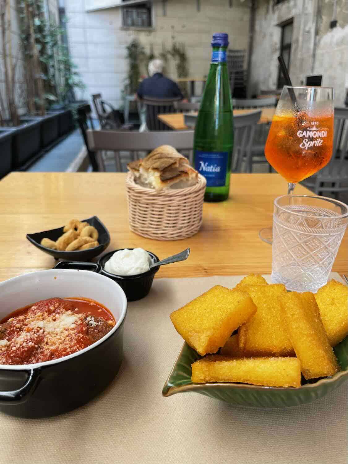 A table set with an assortment of Italian foods, including a dish of meatballs in tomato sauce, a plate of fried polenta sticks, a small bowl of cream, and a basket of bread. A glass of Aperol Spritz and a bottle of sparkling water are also on the table, with an outdoor café setting in the background.