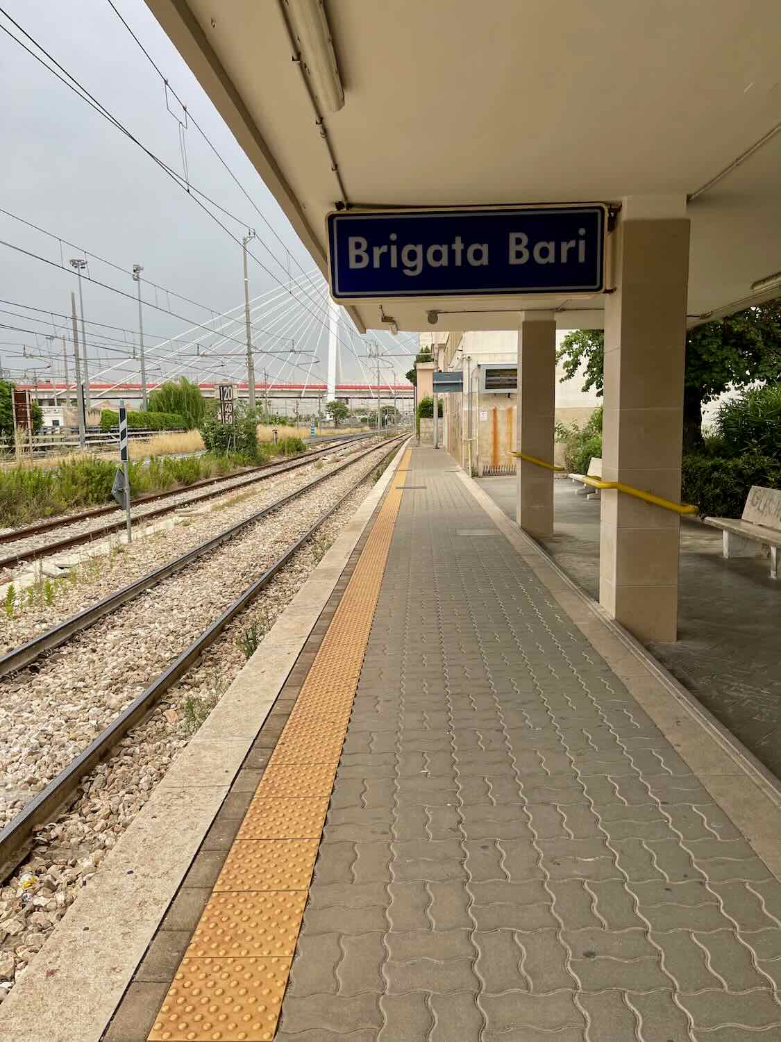 A photo of the Brigata Bari train platform in Bari, Italy. The platform is empty, with tracks running alongside and a sign indicating 'Brigata Bari'. The area is covered with a canopy, and the ground is paved with tiles, including a tactile strip for visually impaired passengers.