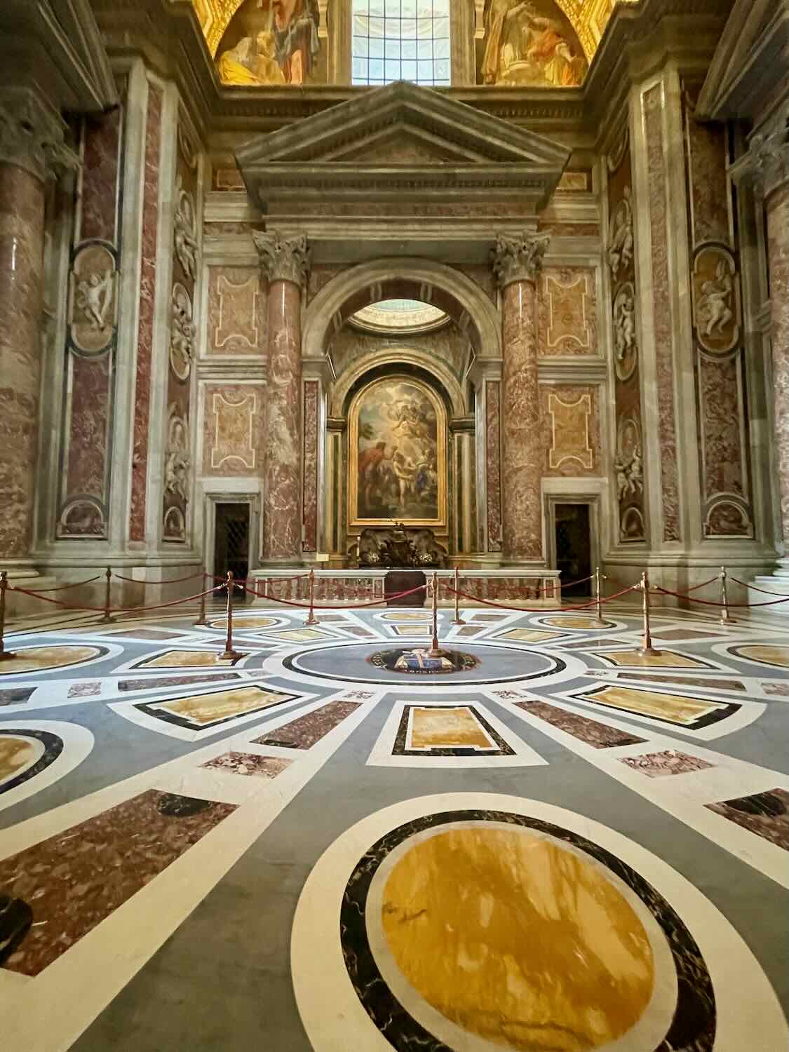 An interior view of a grand room in the Vatican, showcasing an intricate marble floor with circular patterns, tall marble columns, and an ornate altar with religious artwork and statues.