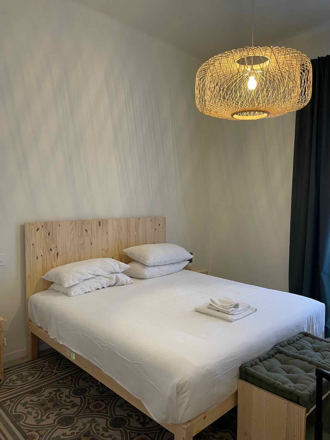 A neatly made bed with white linens in a room at Lostabile Hotel in Bari, Italy. The room has a minimalist design with a woven light fixture hanging from the ceiling and patterned floor tiles.