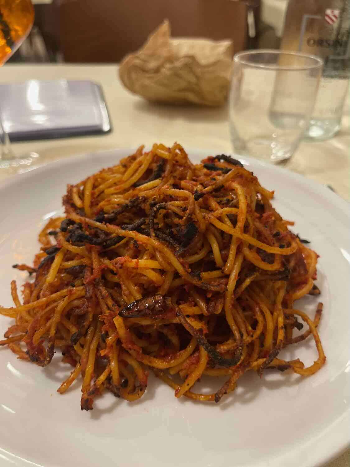 A plate of pasta with a rich tomato-based sauce served in Bari, Italy. The pasta is arranged in a heap on a white plate, with a glass and a bottle of water visible in the background.