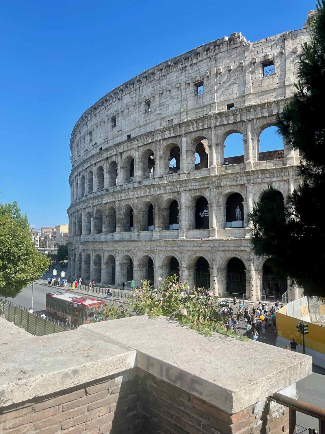 A photo of the Colosseum in Rome on a sunny day, showcasing its ancient, arched structure. The foreground includes some trees and a bus, with tourists visible around the iconic landmark.