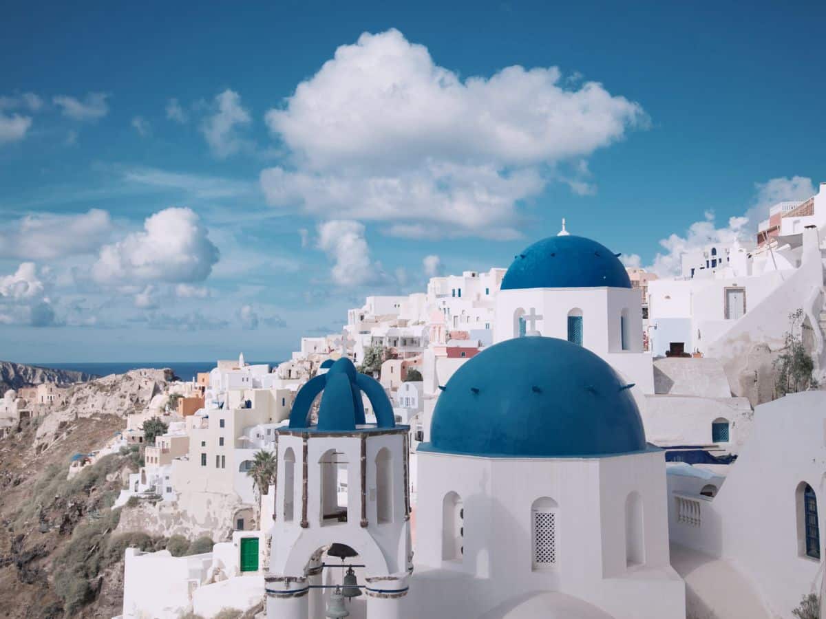 A picturesque scene of whitewashed buildings with blue domes in Santorini, Greece. The buildings are perched on a hillside overlooking the sea, with a bright blue sky and scattered clouds above.