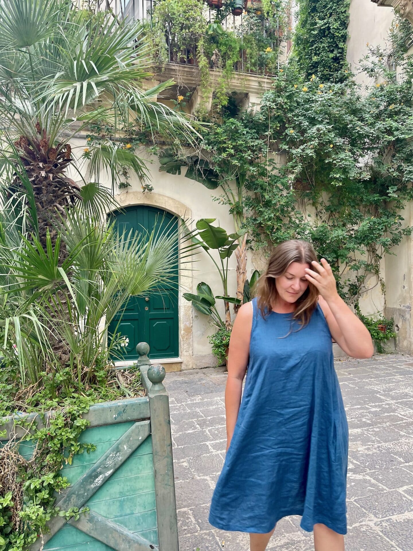 A solo woman in a blue dress stands in front of a green door surrounded by lush greenery in Bari, Italy. She is looking down and touching her hair, with a serene expression on her face.