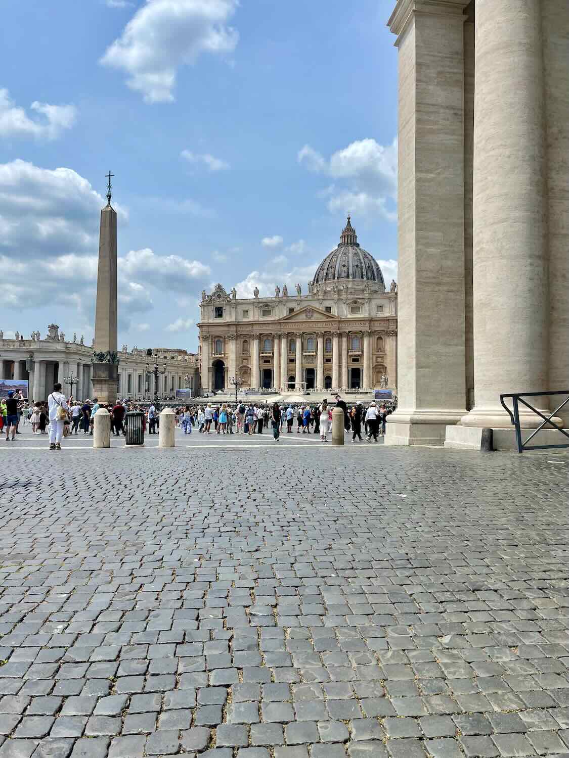 A view of St. Peter's Basilica in Vatican City from St. Peter's Square, with the obelisk in the foreground. The square is bustling with tourists, and the sky is partly cloudy.
