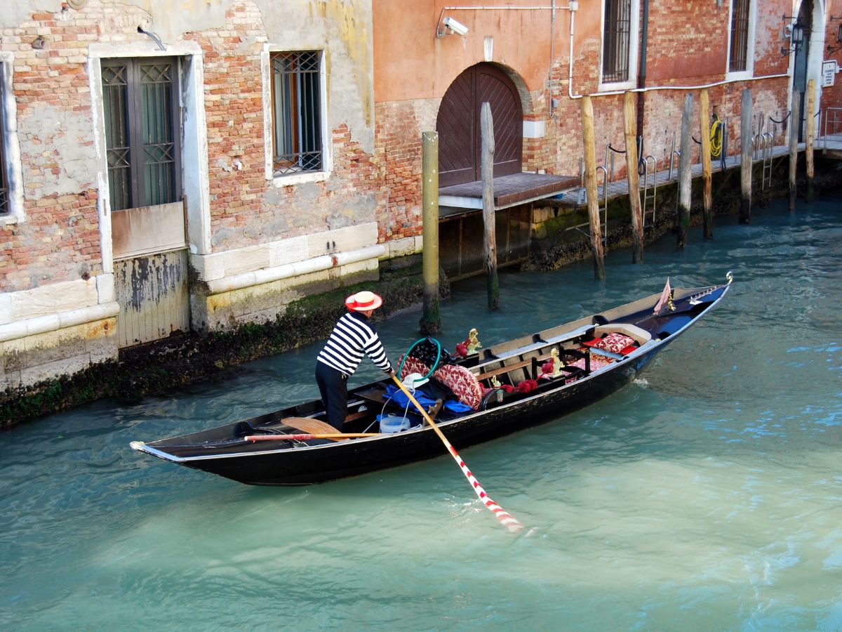 A gondolier in a striped shirt and straw hat navigates a gondola through a narrow canal in Venice, Italy. The gondola passes by old brick buildings with weathered walls and water-stained facades.