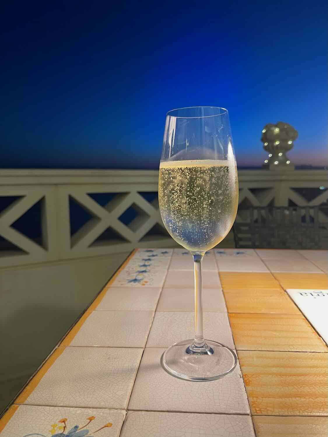A photo of a glass of sparkling wine on a tiled table at dusk. The background features a scenic view of a blue sky transitioning to night, with decorative elements and soft lighting creating an elegant ambiance.