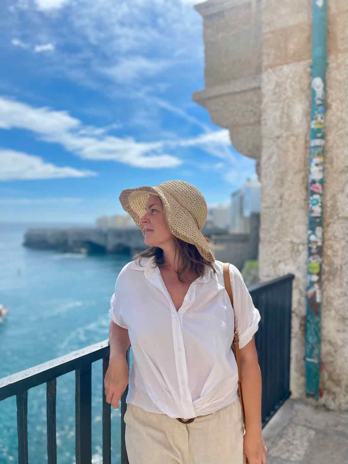 A woman wearing a straw hat and a white shirt stands on a balcony overlooking the sea in Polignano a Mare, Italy. The blue sky and ocean provide a beautiful backdrop as she gazes into the distance.
