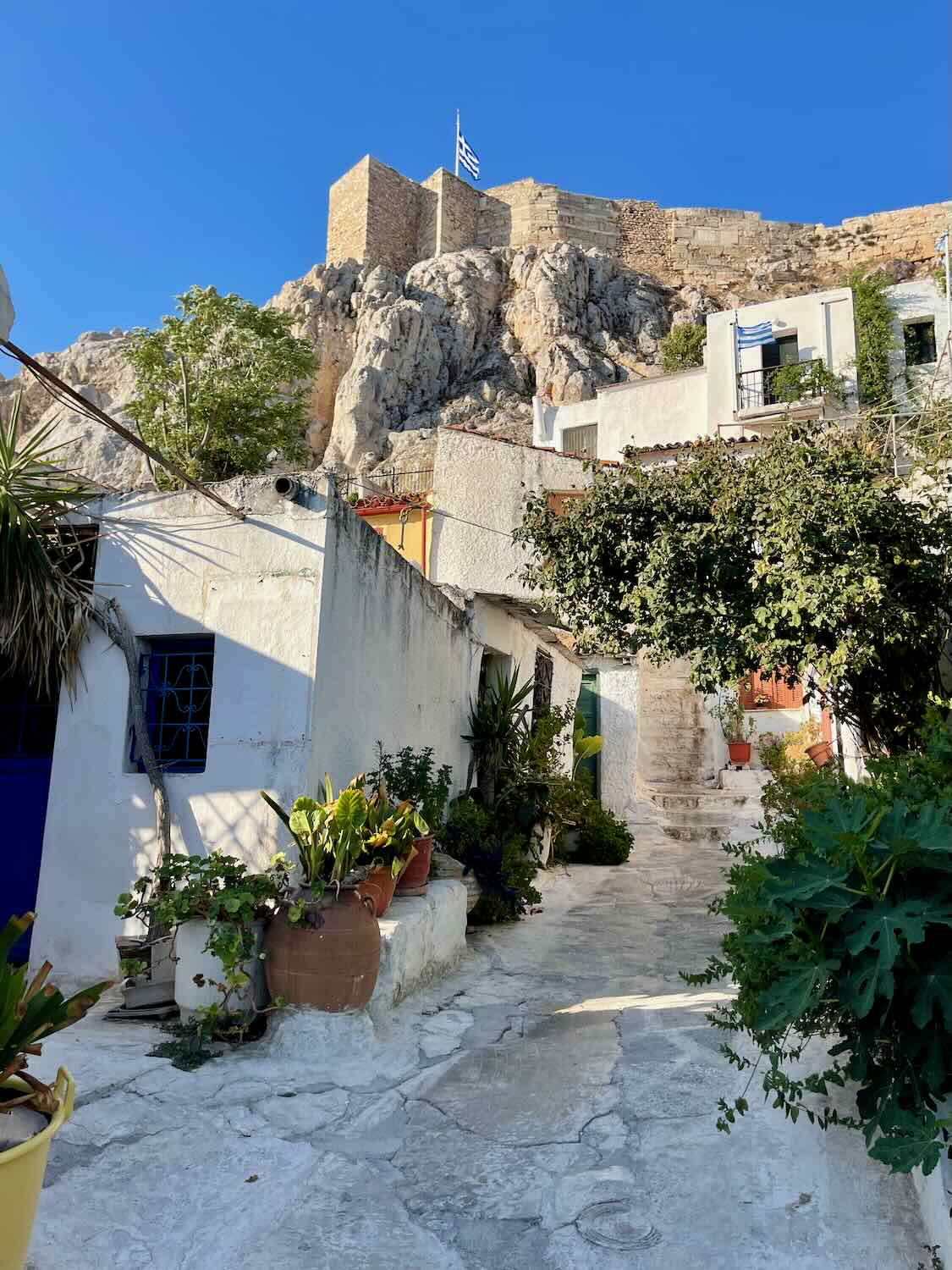 A charming street in the Plaka neighborhood of Athens, featuring white-washed buildings, lush greenery, and the Acropolis with a Greek flag visible in the background.