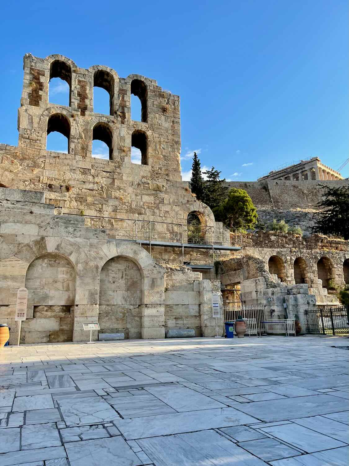 A photo of the Odeon of Herodes Atticus, a stone theatre structure with arched windows and a large stone-paved foreground, located at the Acropolis of Athens, Greece.