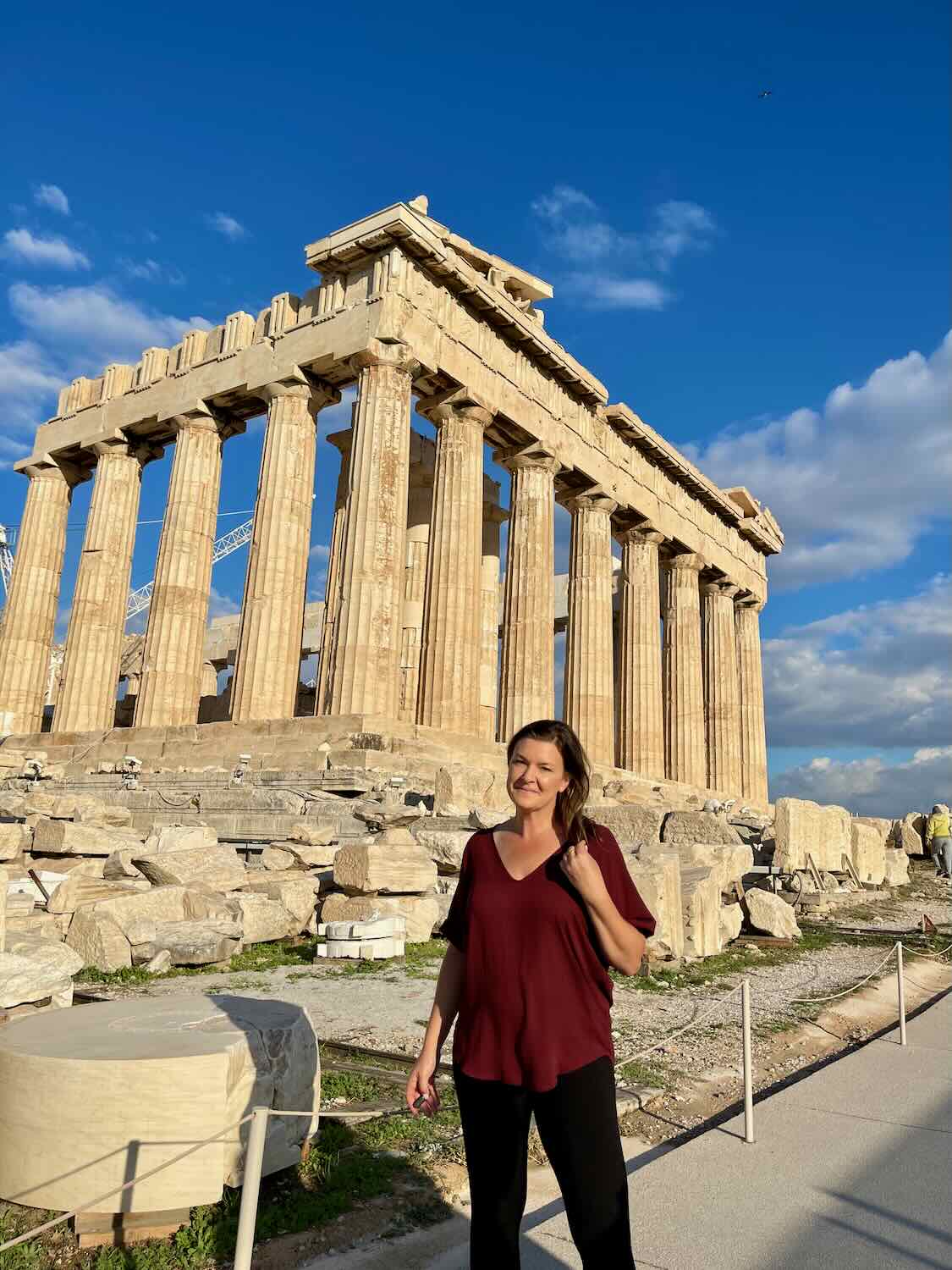 A woman in a red shirt and black pants standing in front of the Parthenon, an ancient Greek temple with iconic Doric columns on the Acropolis of Athens, Greece.