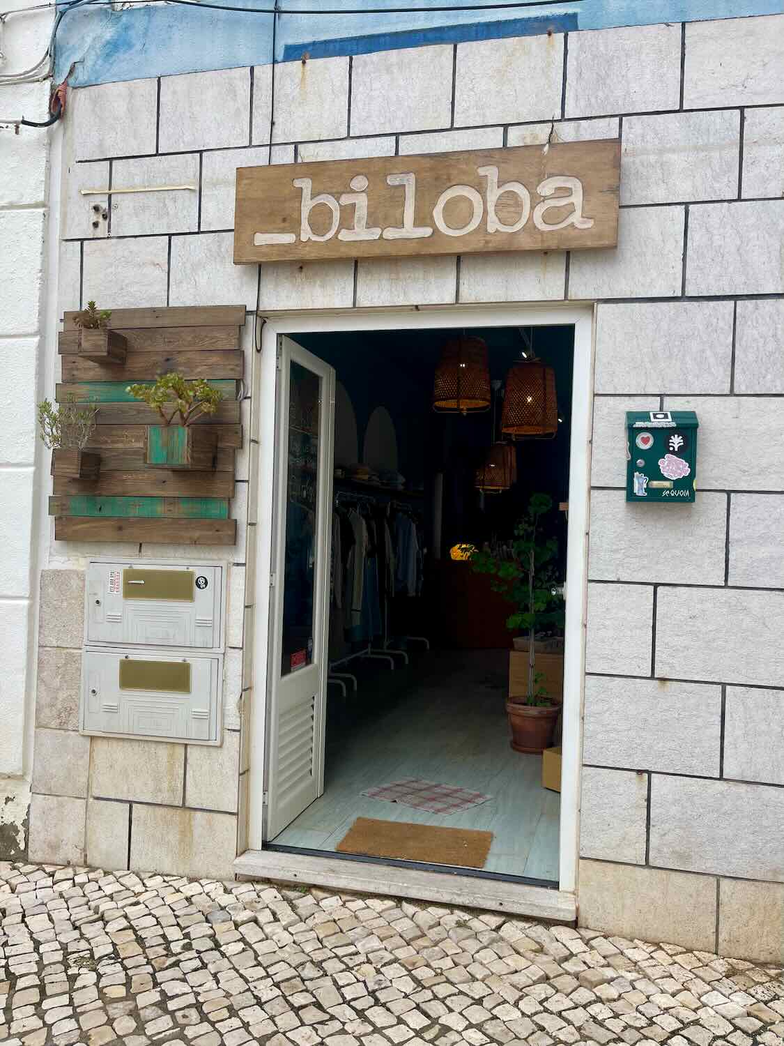 Exterior view of a store named 'Biloba' with a wooden sign above the entrance. The store has a white stone facade with a small plant display to the left of the door. The door is open, revealing the store's interior with warm lighting and a welcoming atmosphere.