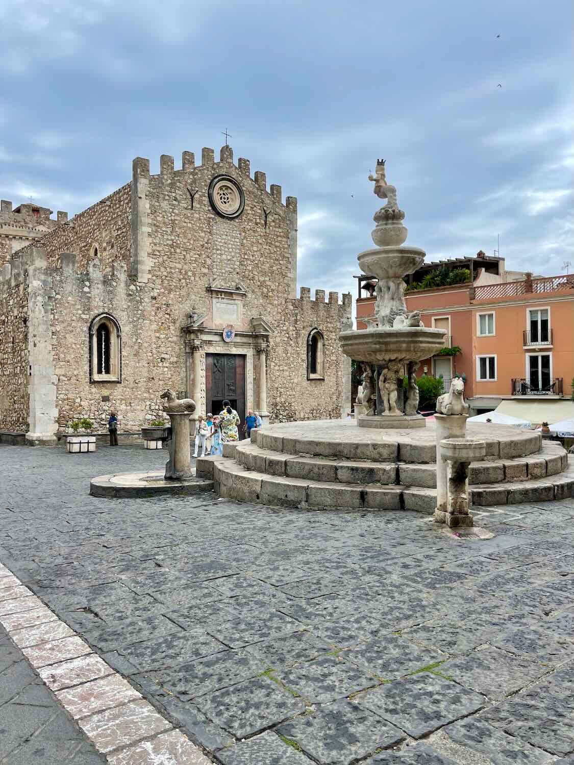 A historic stone church with crenellations stands in a square in Taormina, Italy. In front of the church is a multi-tiered stone fountain with sculptures, including a figure on top. The square is paved with cobblestones, and people are visible near the entrance of the church.