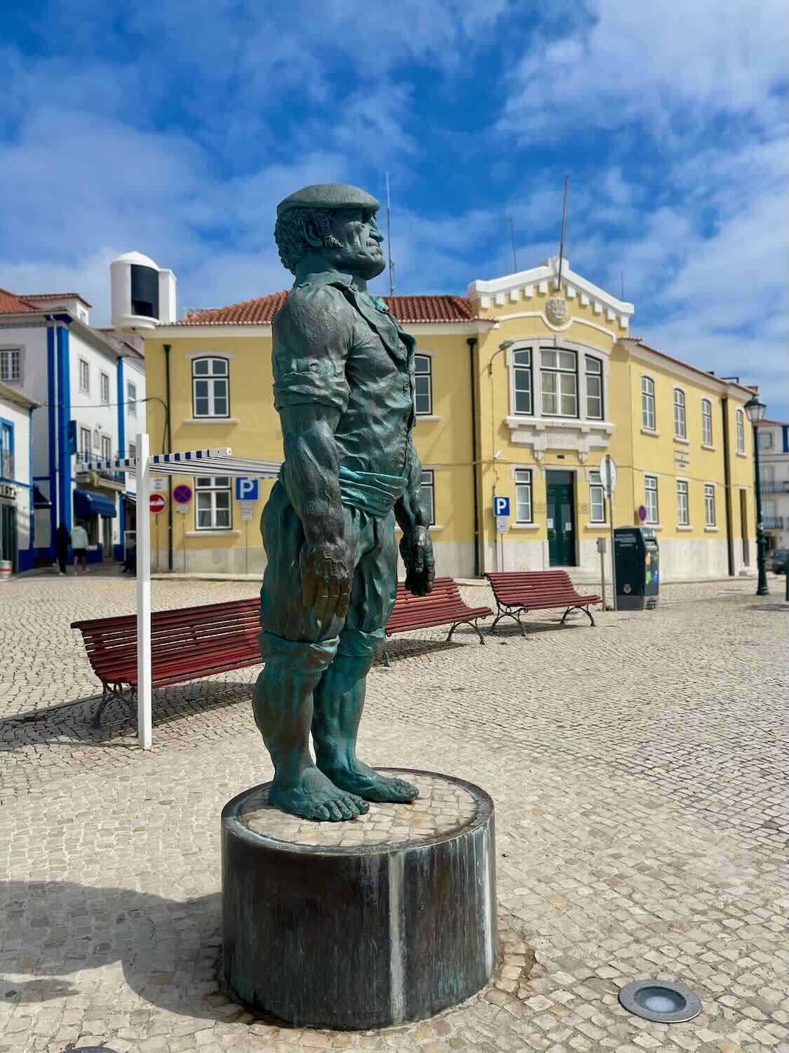 A statue of a robust man standing on a pedestal in a charming town square. The background includes colorful buildings with tiled roofs and a clear blue sky.