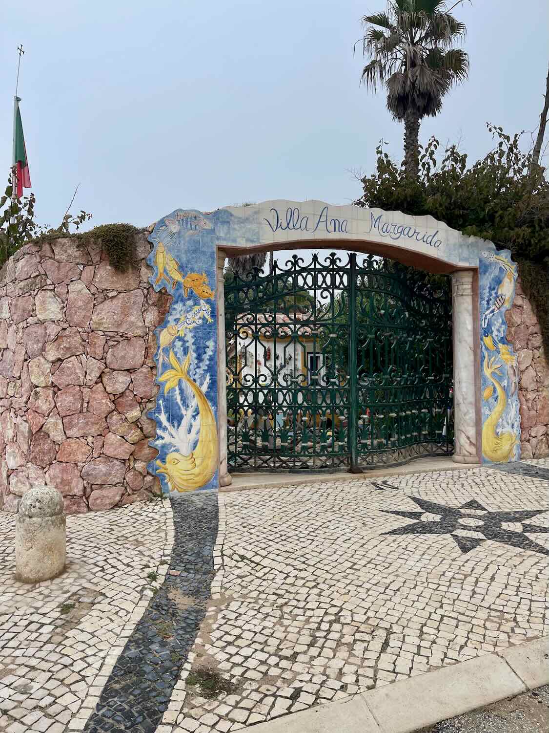 An artistic entrance gate to Villa Ana Margarida in Ericeira. The gate is adorned with colorful murals of fish and aquatic motifs, surrounded by a stone wall and lush greenery.