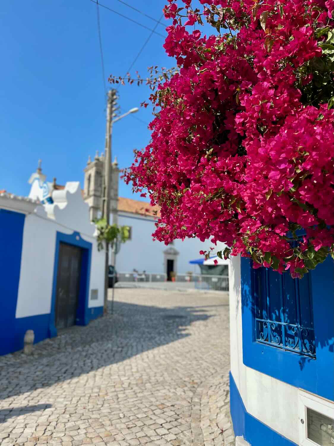 A picturesque street in a quaint town, showcasing white buildings with blue accents and vibrant pink bougainvillea flowers cascading from a wall. The cobblestone path adds to the charm of the scene.