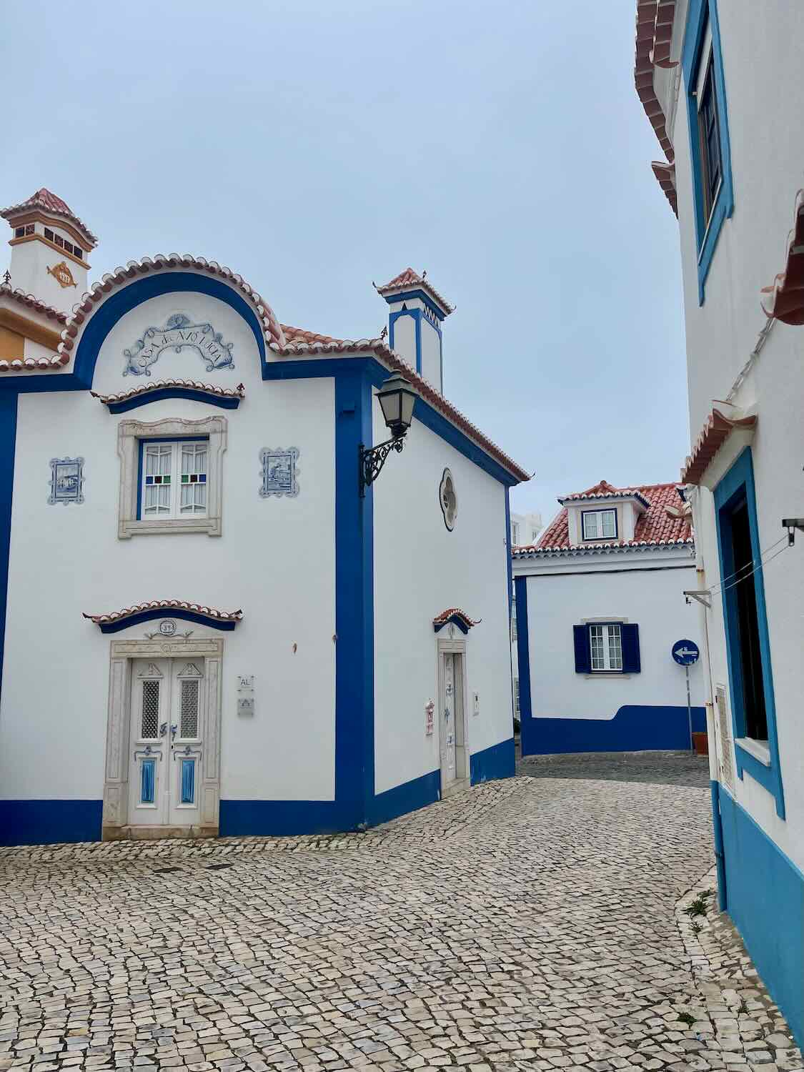 A charming street scene in Ericeira featuring white buildings with blue trim and cobblestone streets. The architecture showcases traditional Portuguese design with red-tiled roofs.