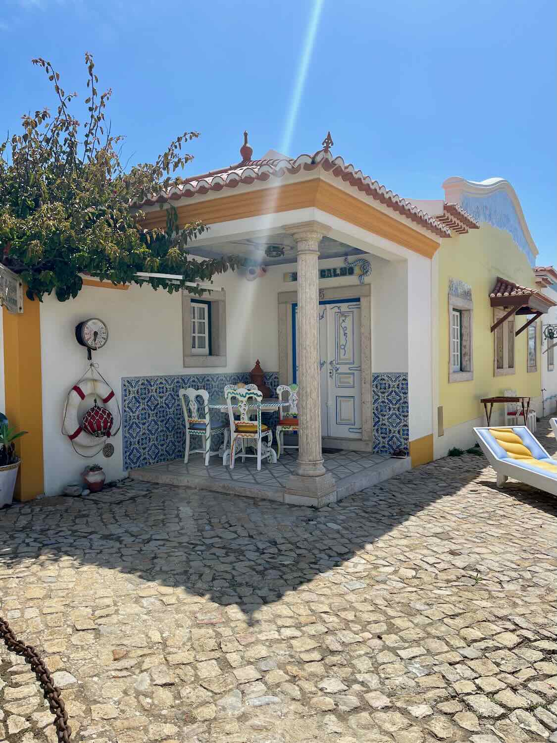 A quaint courtyard of Villa Ana Margarida in Ericeira, with a small dining area featuring a table and chairs. The walls are decorated with blue and white tiles, and a lifebuoy hangs on the wall.