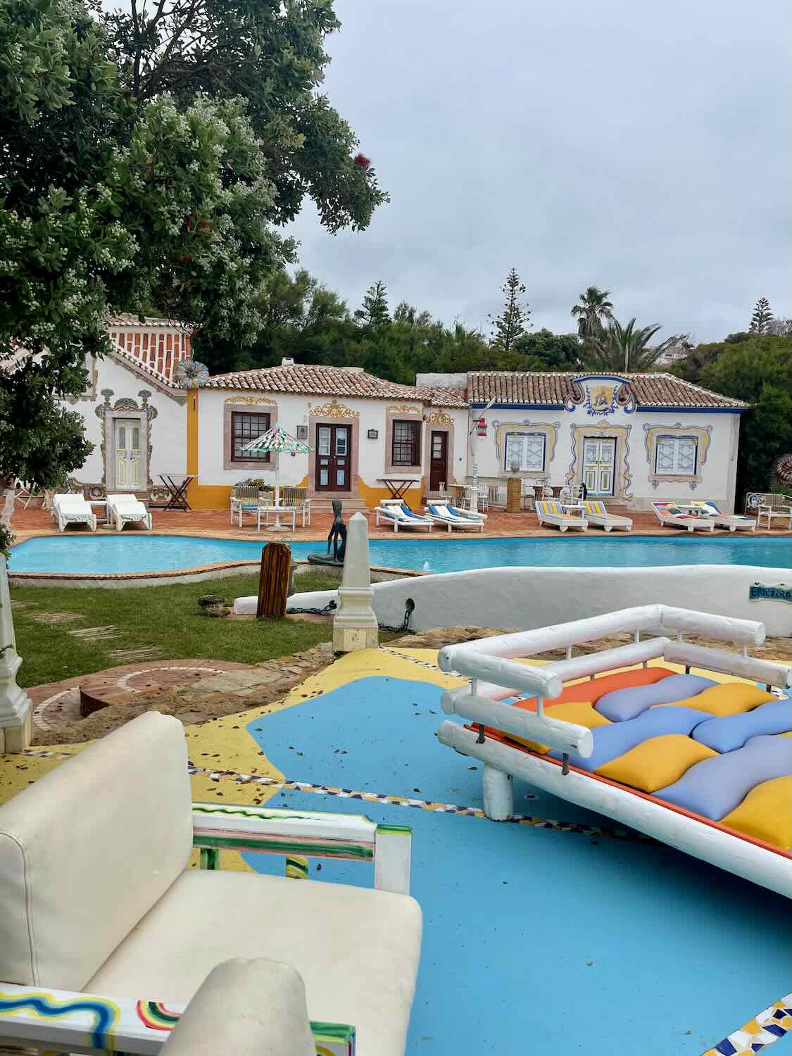 The pool area at Villa Ana Margarida in Ericeira, surrounded by colorful, traditional Portuguese cottages. The scene includes lounge chairs, vibrant decor, and lush greenery.