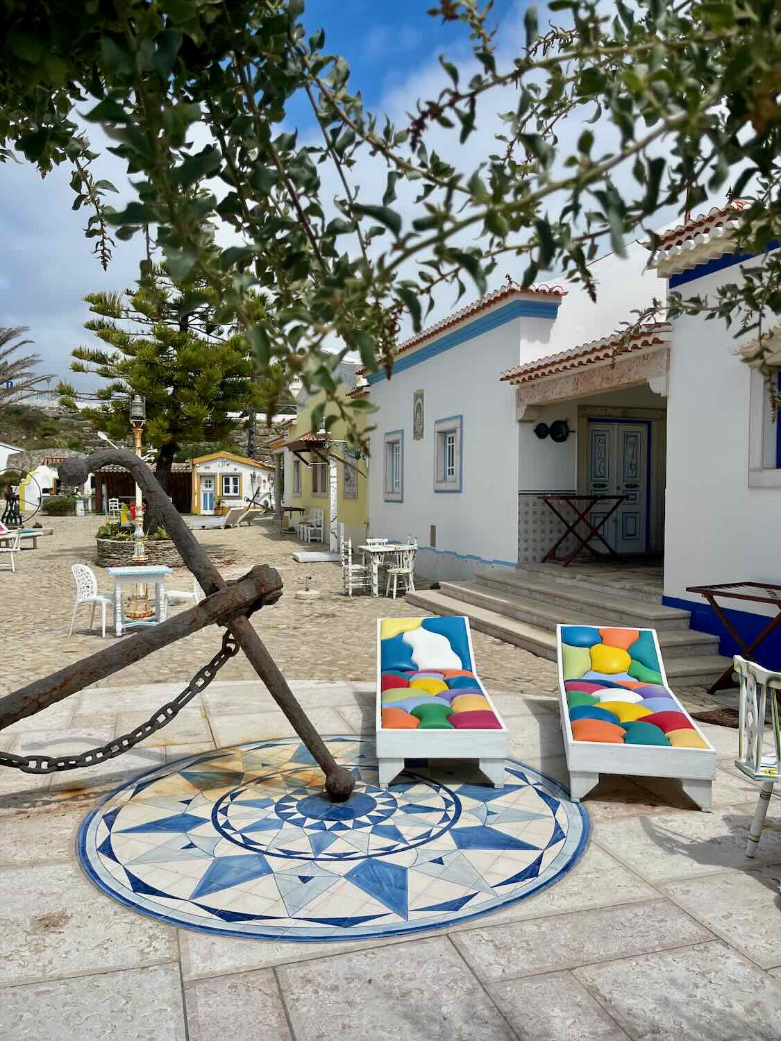 A peaceful courtyard at Villa Ana Margarida in Ericeira, with colorful sun loungers and a large anchor placed on a tiled mosaic. The area is framed by charming cottages and greenery.