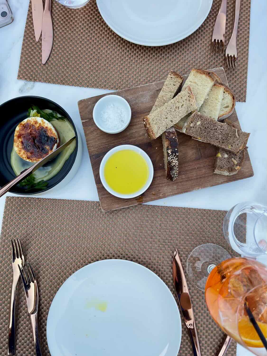 A table setting with plates, cutlery, and a wooden board featuring slices of bread, a bowl of olive oil, and a small dish of salt. A dish with melted cheese and a glass of orange-colored drink are also visible.