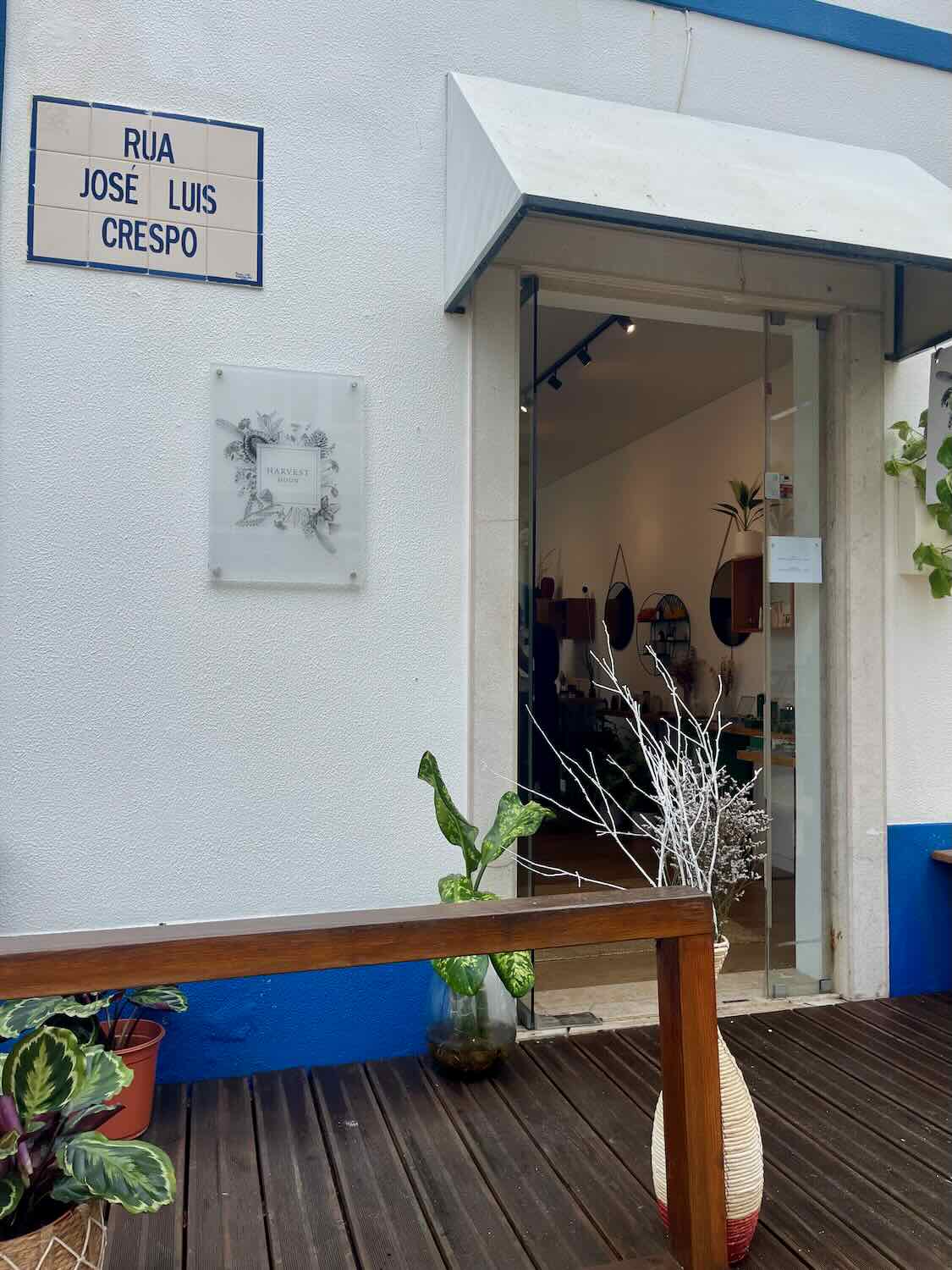 Exterior view of a store named 'Harvest Moon' located on Rua José Luis Crespo. The white storefront features a small awning above the entrance and a sign with botanical illustrations. Plants in pots decorate the entrance, and a wooden bench is placed outside, creating a charming and inviting appearance.