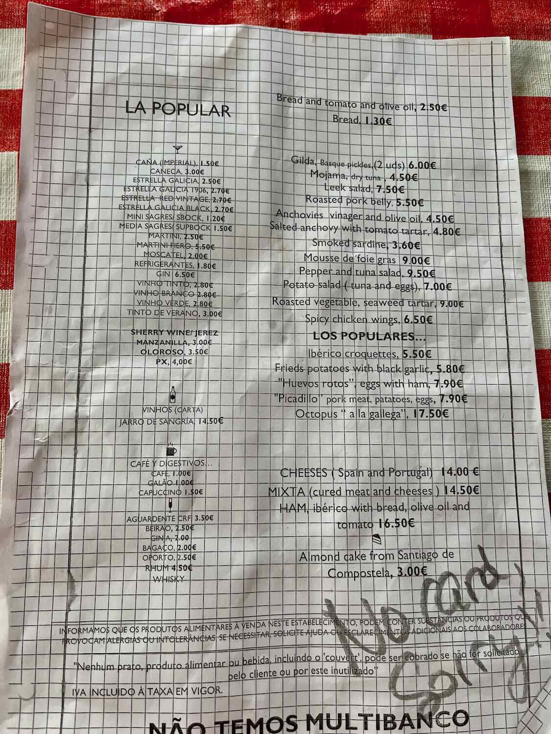 A menu from La Popular, showcasing a variety of items including bread and tomato with olive oil, Gilda Basque pickles, mojama tuna, roasted pork belly, smoked sardine, and various beverages. The menu is placed on a red-checkered tablecloth.