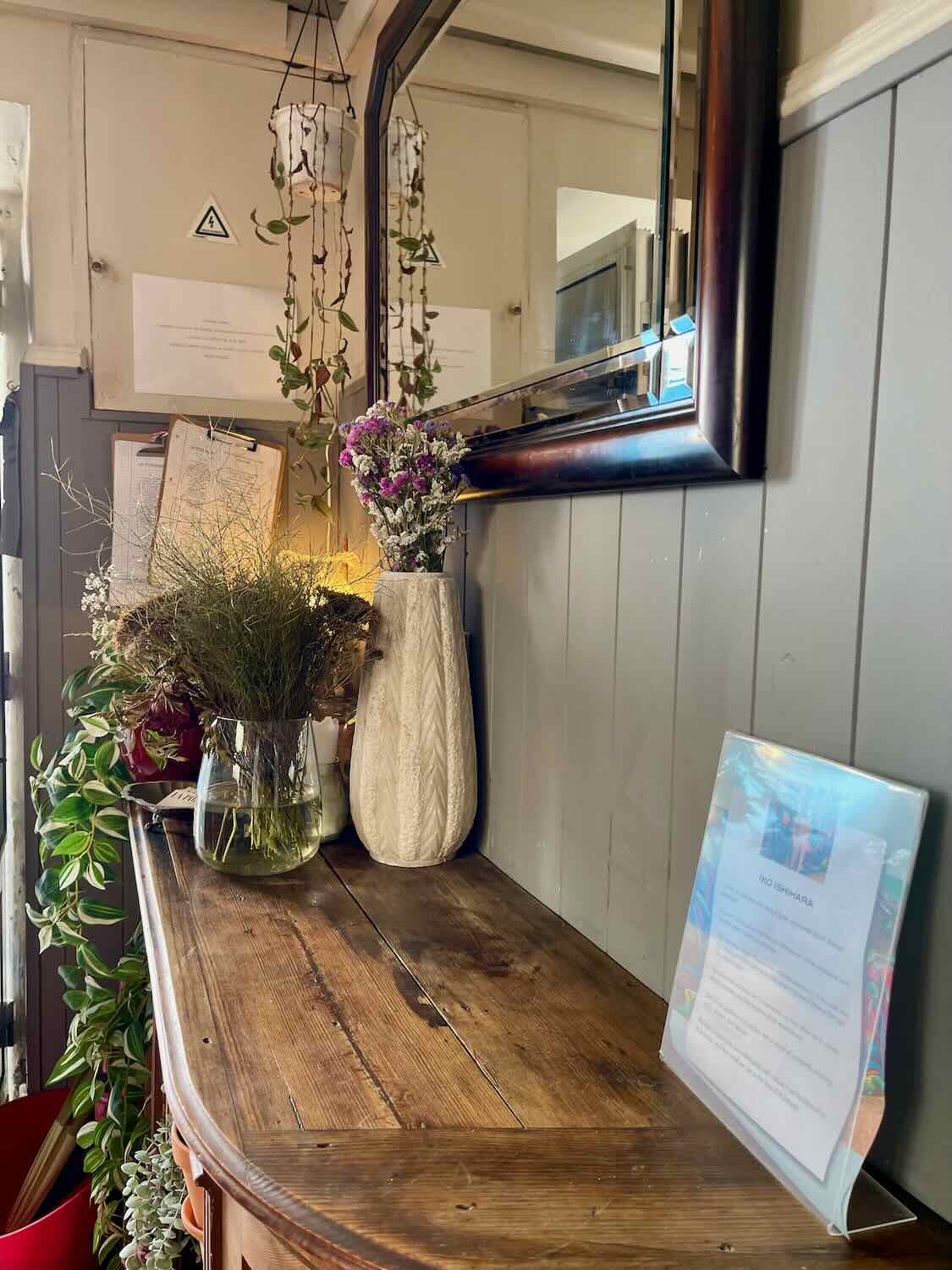 A charming interior setup with a wooden sideboard displaying vases filled with dried and fresh flowers. A large mirror hangs above, reflecting the room and adding depth to the space.