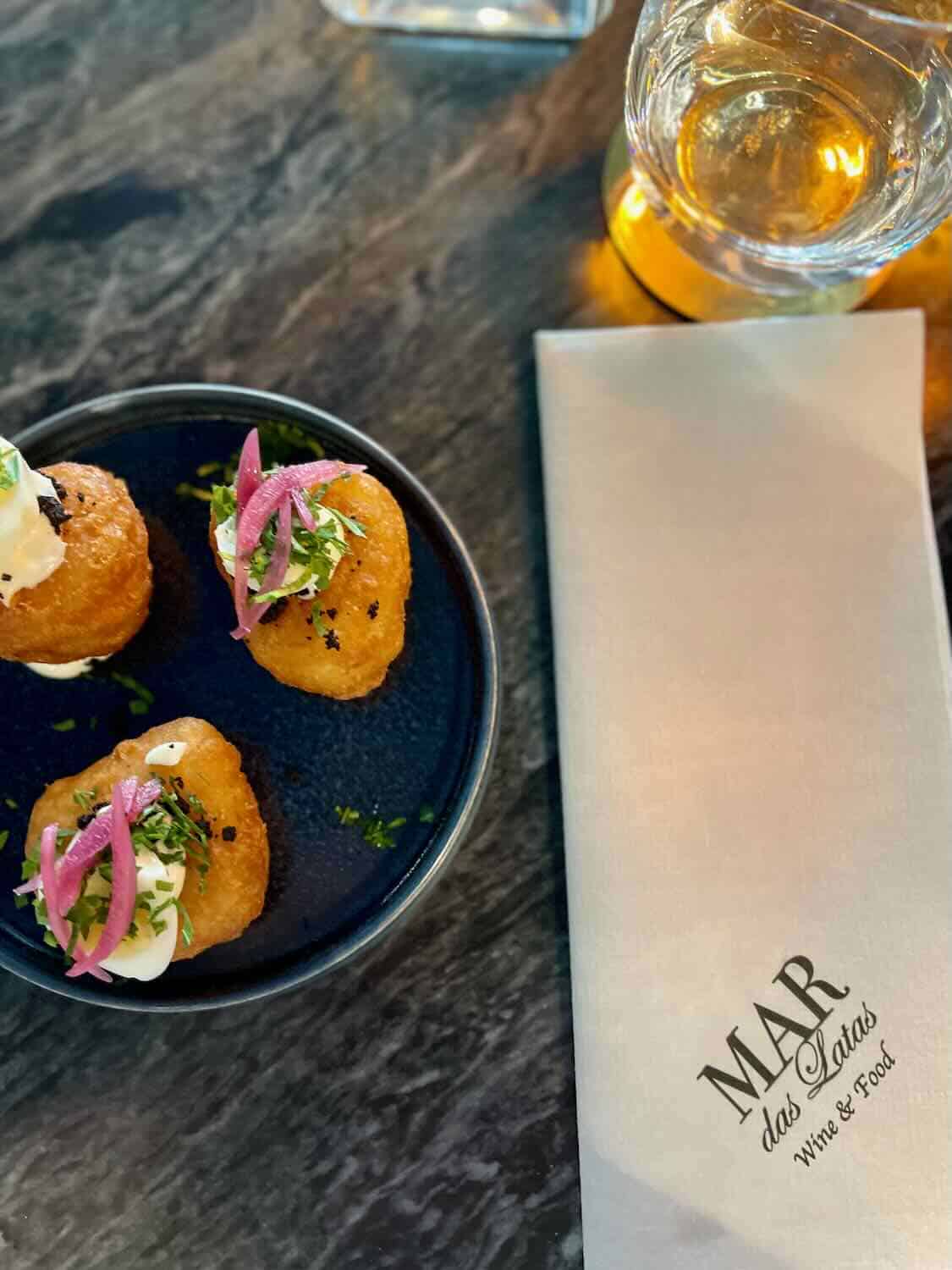 A plate of three small fried appetizers topped with cream and pickled onions, served at Mar das Latas Wine & Food. A glass of white wine and a napkin with the restaurant's logo are next to the plate.