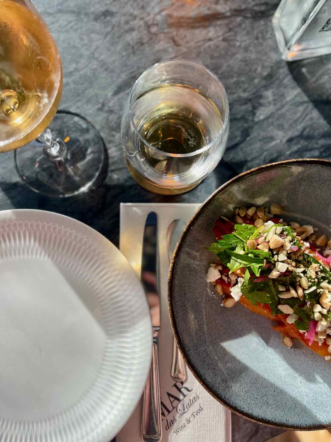 A dining setup at Mar das Latas Wine & Food featuring a bowl of salad topped with nuts, a glass of white wine, and a water glass. The table is set with a white plate and a napkin with the restaurant's logo.