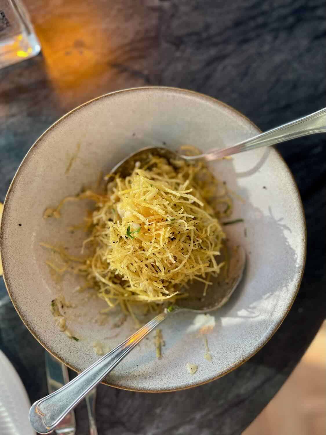 A bowl of finely shredded fried potatoes served at Mar das Latas Wine & Food, placed on a dark marble table. The bowl is accompanied by a serving spoon.