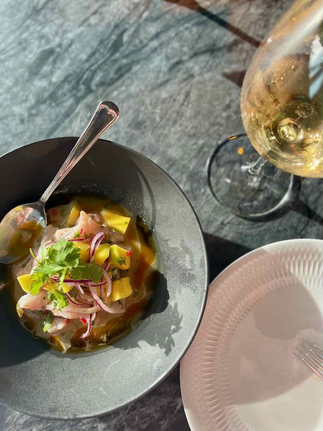 A bowl of ceviche garnished with cilantro and red onions, served at Mar das Latas Wine & Food. A glass of white wine and a white plate are set beside the bowl on a dark marble table.