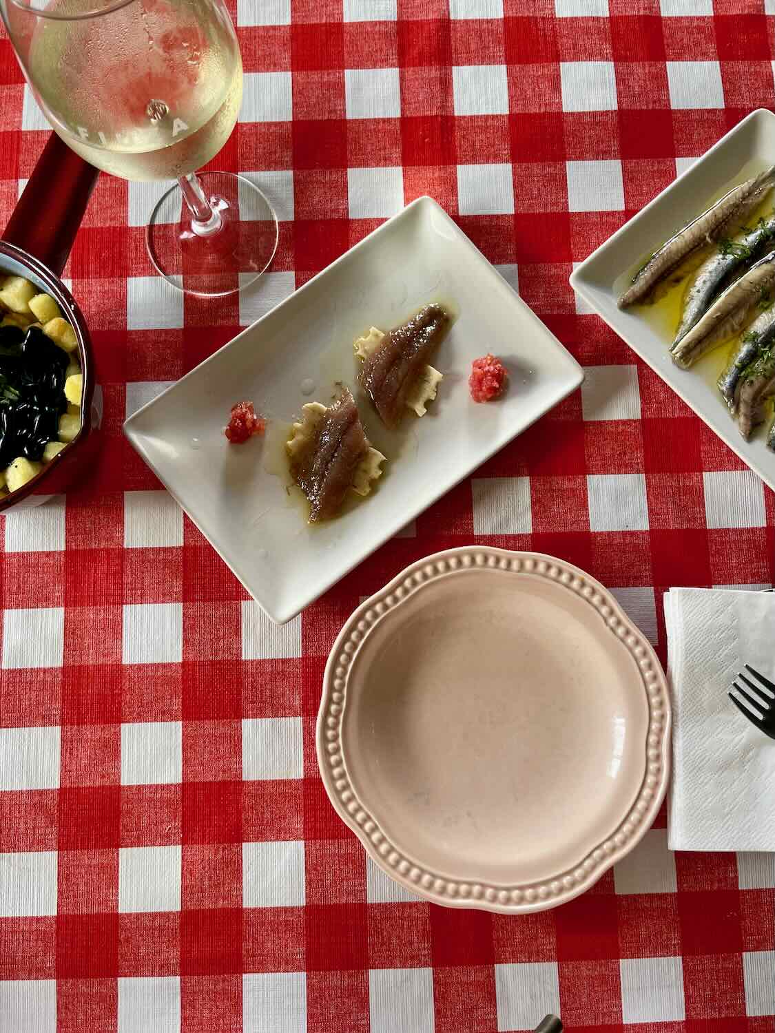 A dining setup on a red-checkered tablecloth featuring plates of anchovies and a glass of white wine. The table also includes a dish of olives and a pink ceramic bowl.