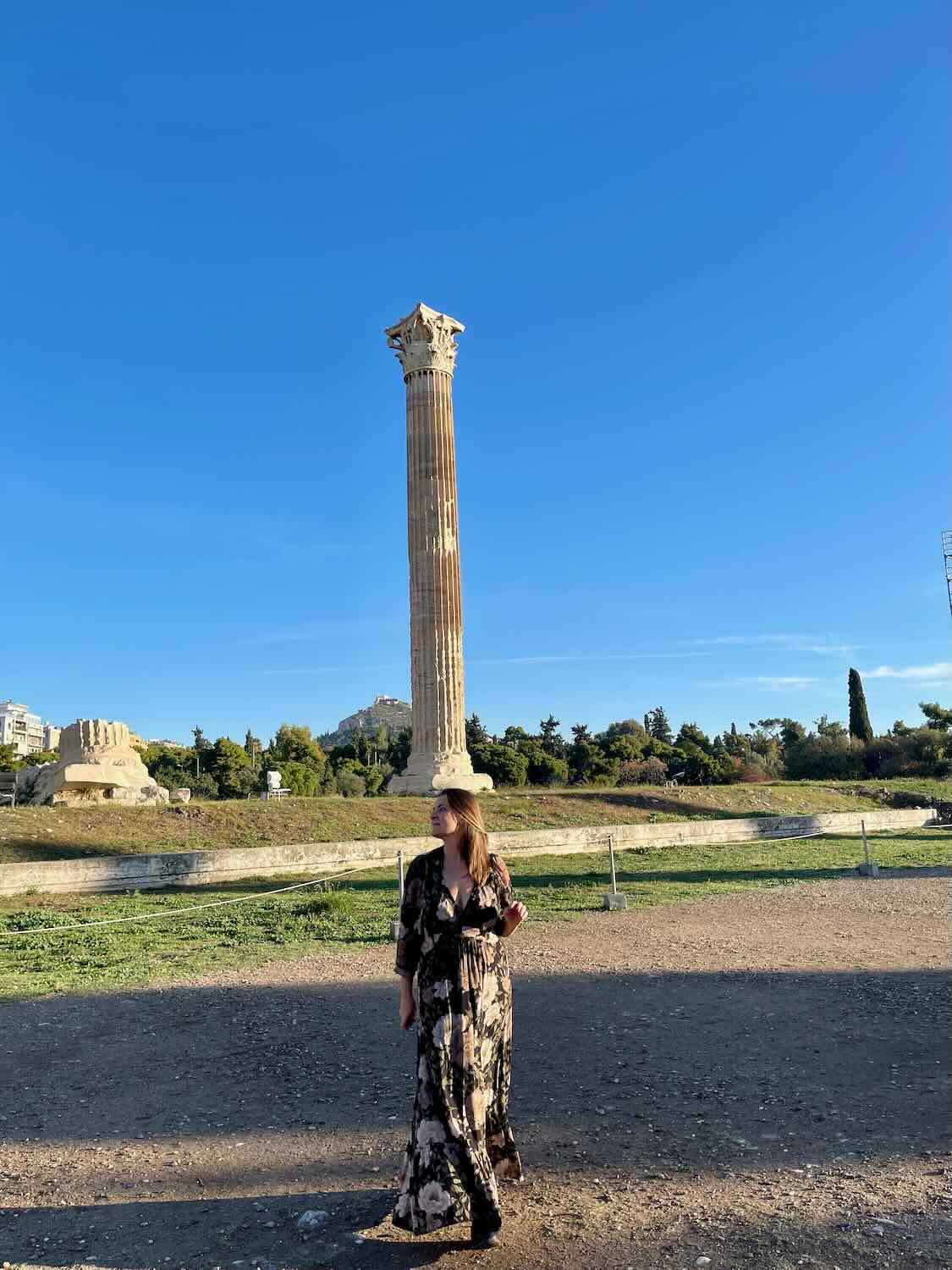 A woman in a floral dress and sun hat standing in front of a single standing column of the Temple of Olympian Zeus in Athens, with a clear blue sky in the background.