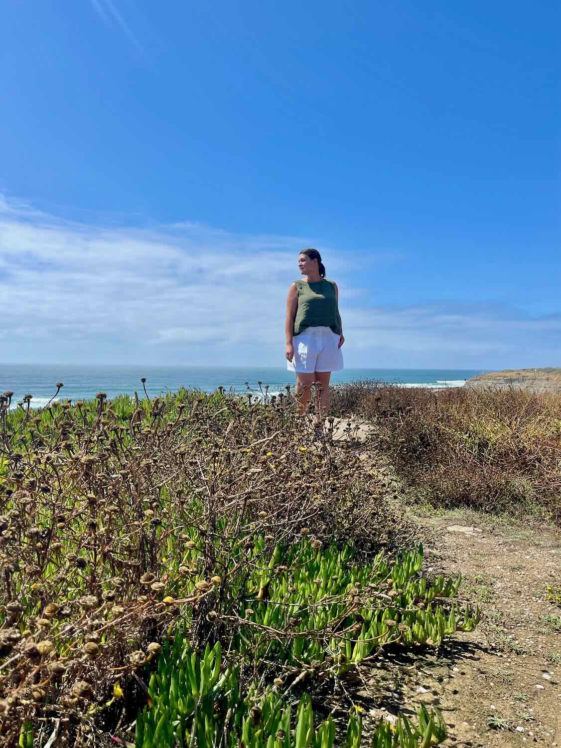 A woman standing on a coastal path with green shrubs, looking out towards the ocean under a clear blue sky. She is wearing a green tank top and white shorts, enjoying the sunny day in Ericeira.