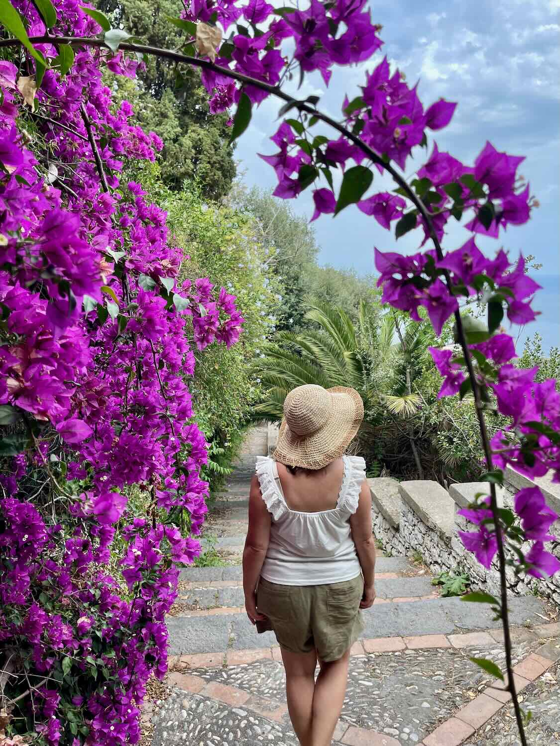 A woman going down the stairs with a view of greenery and purple flowers. 