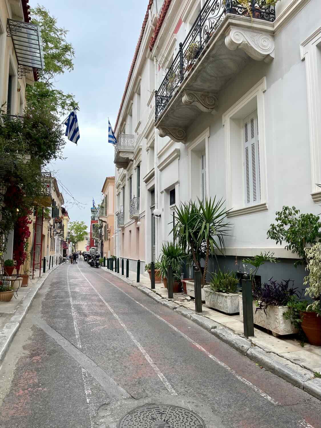 A narrow street in Athens lined with potted plants and classic buildings with balconies. Greek flags are visible, and the street is calm and empty.