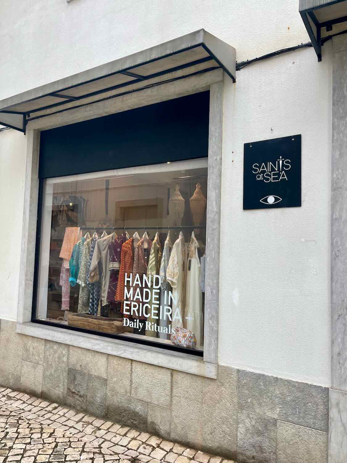 The exterior of a boutique named "Saints at Sea" with a display window showcasing a variety of colorful handmade clothing. The store's sign is visible on the wall next to the window, and the pavement outside is made of cobblestones. The display window highlights the text "Hand Made in Ericeira - Daily Rituals," emphasizing the artisanal nature of the products sold inside.