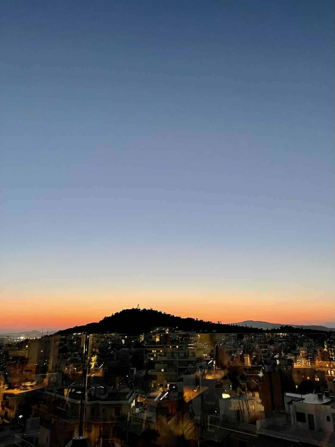 A view of Athens at sunset with Mount Lycabettus silhouetted against a colorful sky transitioning from orange to deep blue.