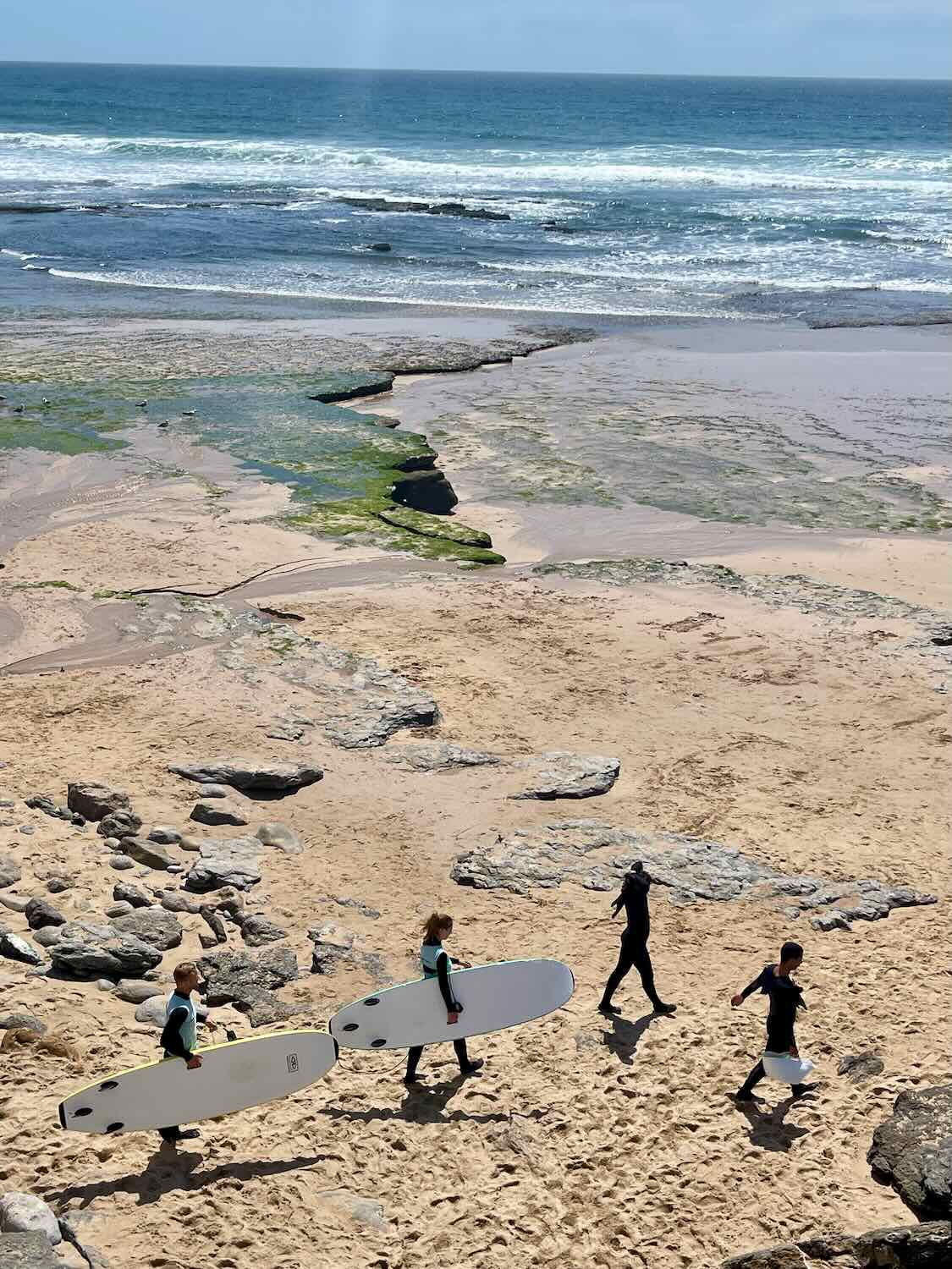 Three surfers walk along a sandy beach in Ericeira, Portugal, carrying surfboards under their arms. The beach is dotted with rocks and patches of green seaweed, with waves gently rolling in the background under a bright, clear sky.