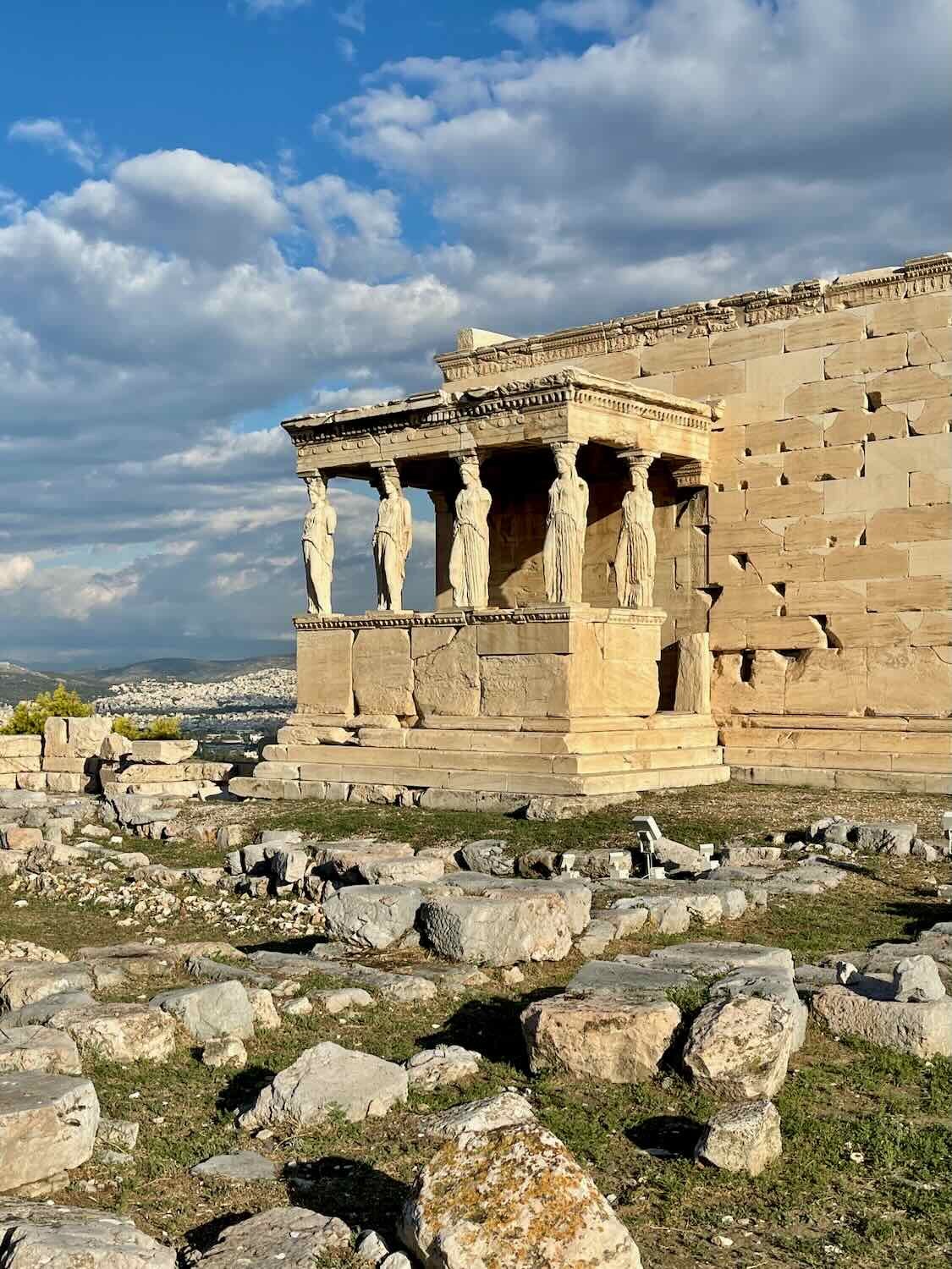 A photo of the Porch of the Caryatids at the Erechtheion, featuring six draped female figures as supporting columns, with a backdrop of the Acropolis and cloudy sky.