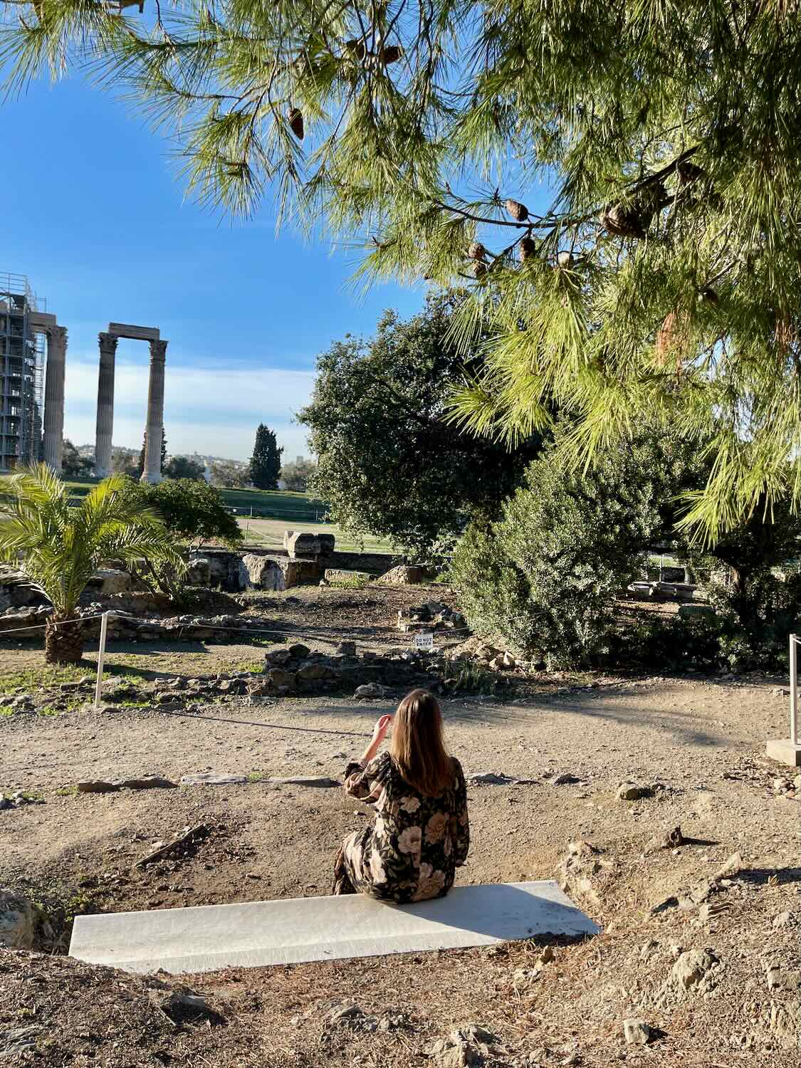 A woman sitting on a stone bench, photographing the ruins of the Temple of Olympian Zeus in Athens. The scene is framed by pine trees and greenery under a clear blue sky.