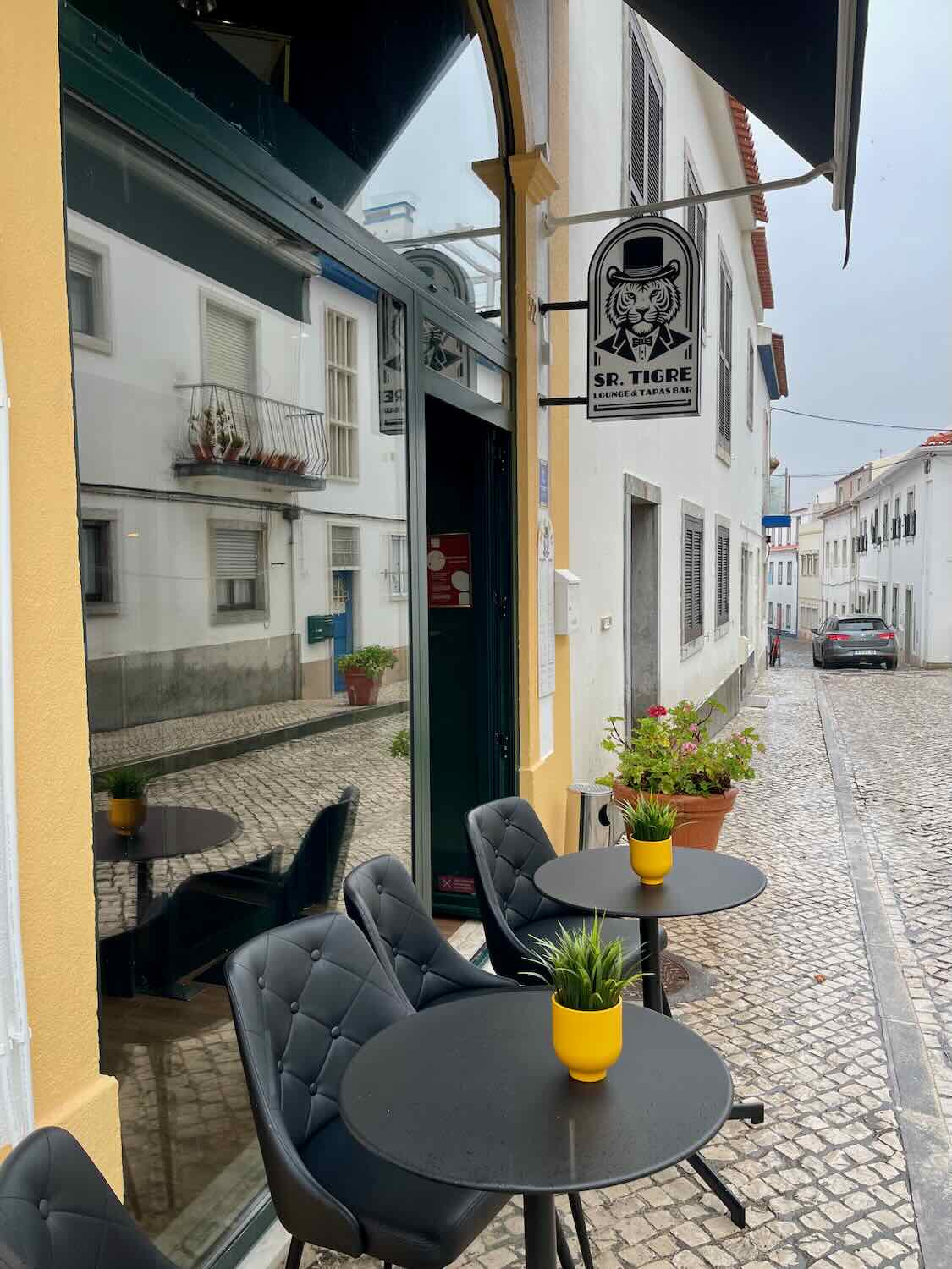 The exterior of Sr. Tigre Lounge & Tapas Bar, featuring a modern sign with a tiger logo and outdoor seating with black tables and chairs. The cobblestone street and surrounding white buildings add to the charming ambiance.